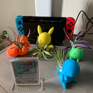 May include: A collection of air plants in 3D printed Pokemon planters. The planters are shaped like Pikachu, Squirtle, and Bulbasaur. The plants are green and brown. There is a Pokemon card in a clear plastic case on the table.
