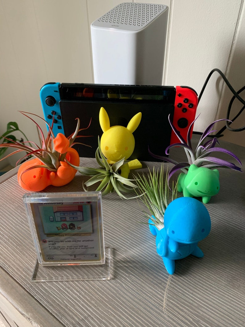 May include: A collection of air plants in 3D printed Pokemon planters. The planters are shaped like Pikachu, Squirtle, and Bulbasaur. The plants are green and brown. There is a Pokemon card in a clear plastic case on the table.