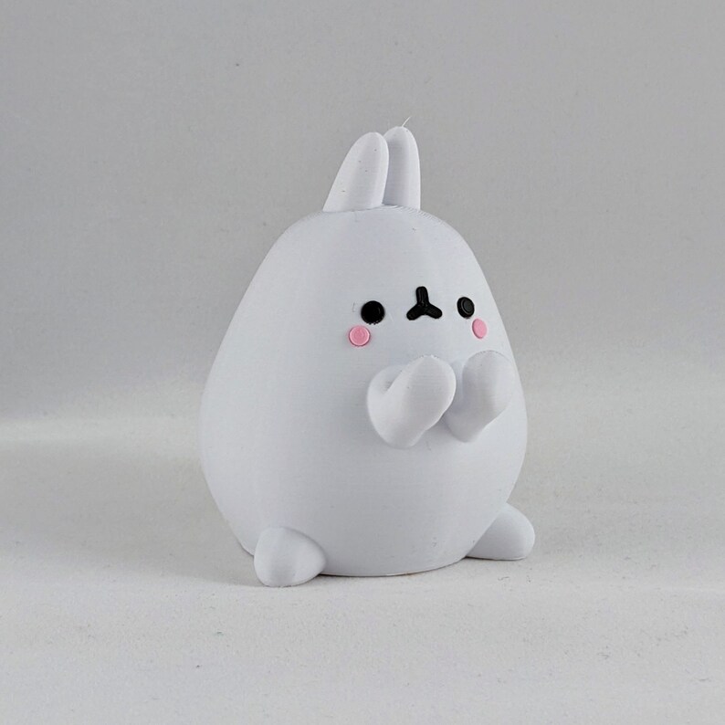 May include: White bunny figurine with pink cheeks and black eyes. The bunny is sitting with its paws raised in front of it.