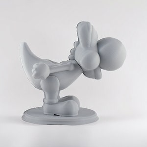 May include: A gray 3D printed figurine of a cartoon character with a long neck, large head, and a long tail. The character is in a crouching position with its head tilted down.