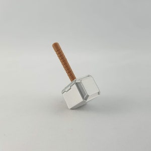 May include: A small, white plastic toy hammer with a brown wooden handle. The hammer is shaped like Thor's hammer, Mjolnir.