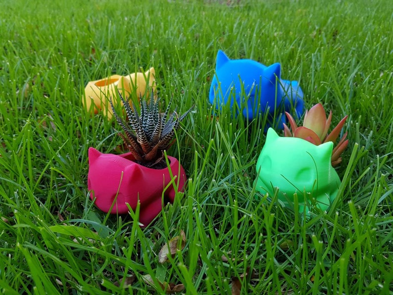 May include: Four colorful ceramic planters shaped like cartoon cats, with two of the planters containing small succulents. The planters are in shades of yellow, red, blue, and green, and are sitting on a bed of green grass.