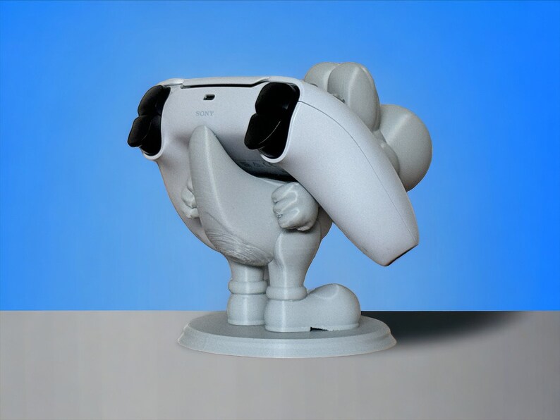 May include: White 3D printed figurine of a cartoon character holding a white PlayStation 5 controller. The character is standing on a round base.