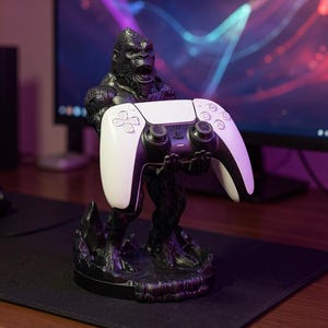 May include: A black gorilla-shaped controller stand holding a white and black game controller. The stand is on a black surface, with a computer monitor in the background. The controller has a sleek, modern design.