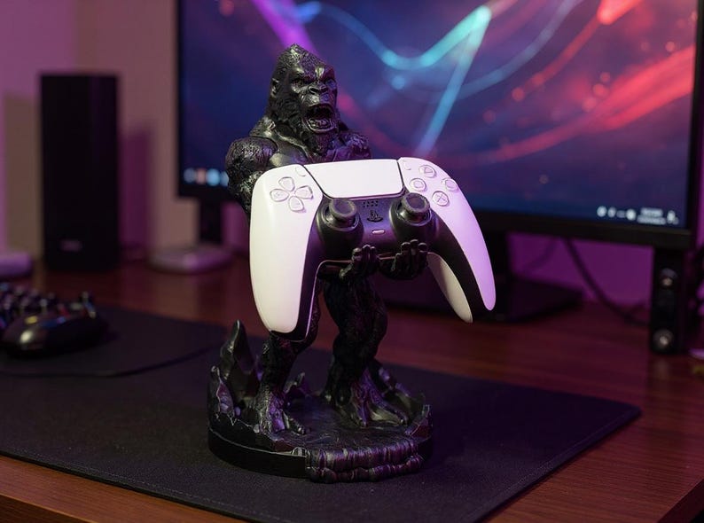 May include: A black gorilla-shaped controller stand holding a white and black game controller. The stand is on a black surface, with a computer monitor in the background. The controller has a sleek, modern design.