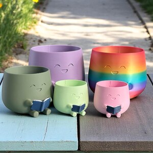 K&ouml;nnte beinhalten: Eine Sammlung von f&uuml;nf bunten Pflanzgef&auml;&szlig;en mit l&auml;chelnden Gesichtern. Die Pflanzgef&auml;&szlig;e sind in Lila-, Gr&uuml;n-, Rosa- und Regenbogenfarben gehalten. Zwei kleinere Pflanzgef&auml;&szlig;e halten blaue B&uuml;cher. Die Pflanzgef&auml;&szlig;e stehen auf einer Holzoberfl&auml;che.