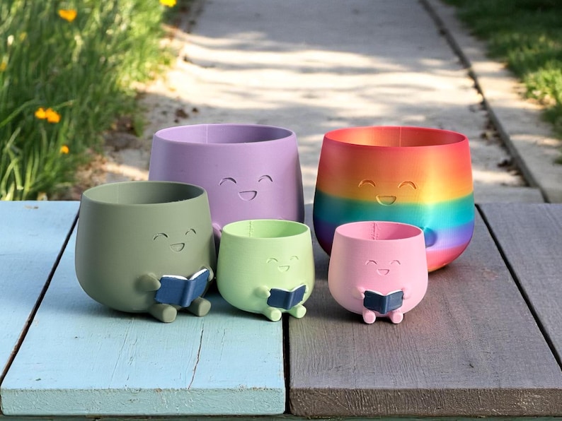 K&ouml;nnte beinhalten: Eine Sammlung von f&uuml;nf bunten Pflanzgef&auml;&szlig;en mit l&auml;chelnden Gesichtern. Die Pflanzgef&auml;&szlig;e sind in Lila-, Gr&uuml;n-, Rosa- und Regenbogenfarben gehalten. Zwei kleinere Pflanzgef&auml;&szlig;e halten blaue B&uuml;cher. Die Pflanzgef&auml;&szlig;e stehen auf einer Holzoberfl&auml;che.