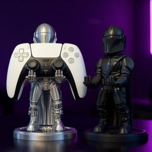 Pode incluir: Dois suportes para comando com o tema Mandalorian. Um &eacute; prateado e segura um comando PlayStation 5 branco e preto. O outro &eacute; preto e apresenta uma figura de Mandalorian. Ambos est&atilde;o em bases redondas.