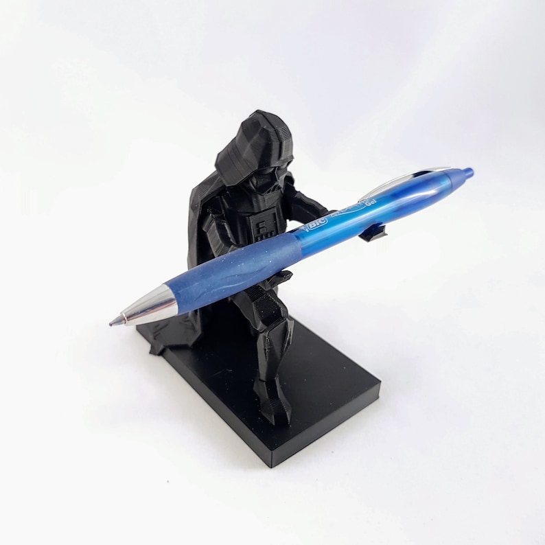 May include: A black 3D-printed Darth Vader pen holder with a blue BIC pen. The pen holder is in a kneeling position, holding the pen. The pen is blue with silver accents. The pen holder sits on a black rectangular base.