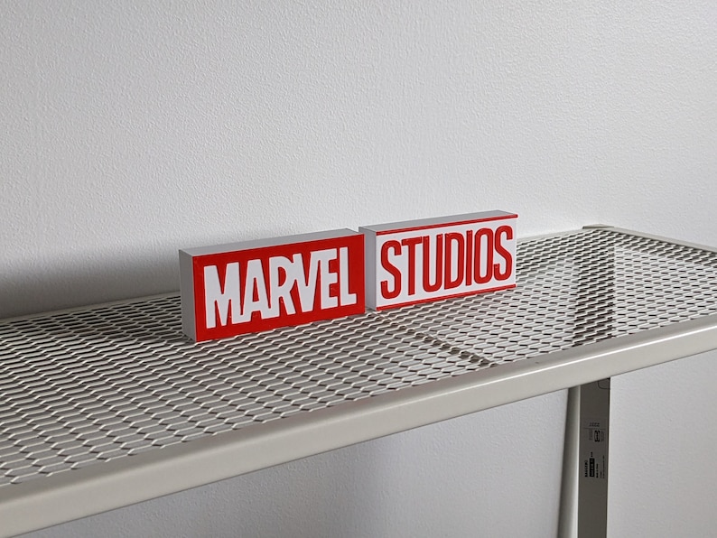 K&ouml;nnte beinhalten: Zwei rote Bl&ouml;cke mit wei&szlig;en Buchstaben auf einem wei&szlig;en Metallregal. Die Bl&ouml;cke zeigen "MARVEL" und "STUDIOS".