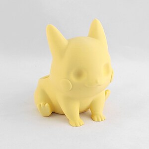 May include: A yellow plastic figurine of a cartoon character with large ears and a rounded body. The figurine is sitting with its legs crossed and its arms resting on its lap.