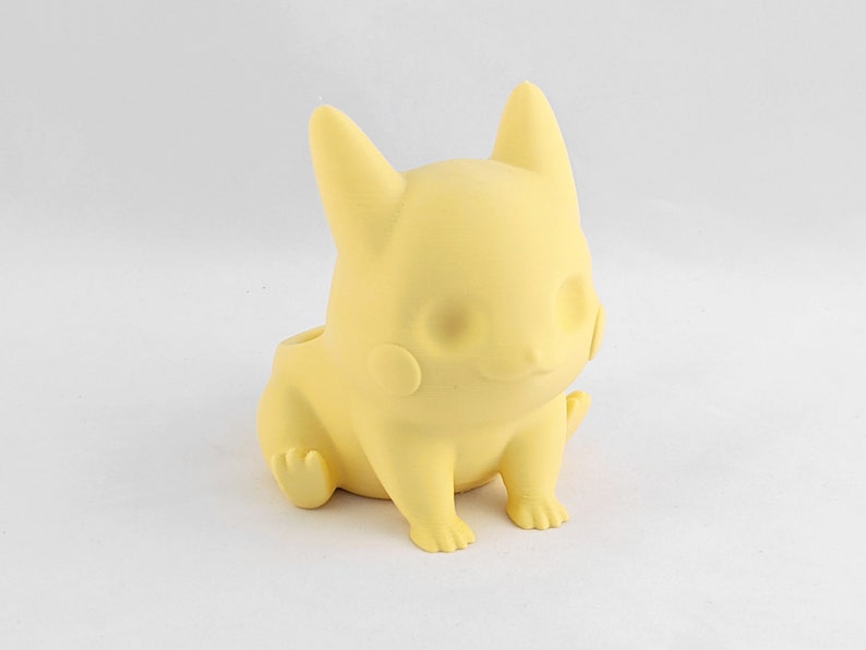 May include: A yellow plastic figurine of a cartoon character with large ears and a rounded body. The figurine is sitting with its legs crossed and its arms resting on its lap.