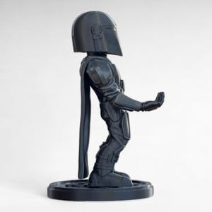 Pode incluir: Figura de a&ccedil;&atilde;o preta de um Mandaloriano, em p&eacute; sobre uma base redonda. A figura est&aacute; em uma pose din&acirc;mica, com um bra&ccedil;o estendido. O capacete e a armadura s&atilde;o detalhados, e uma capa pende nas costas. Feito de material escuro.