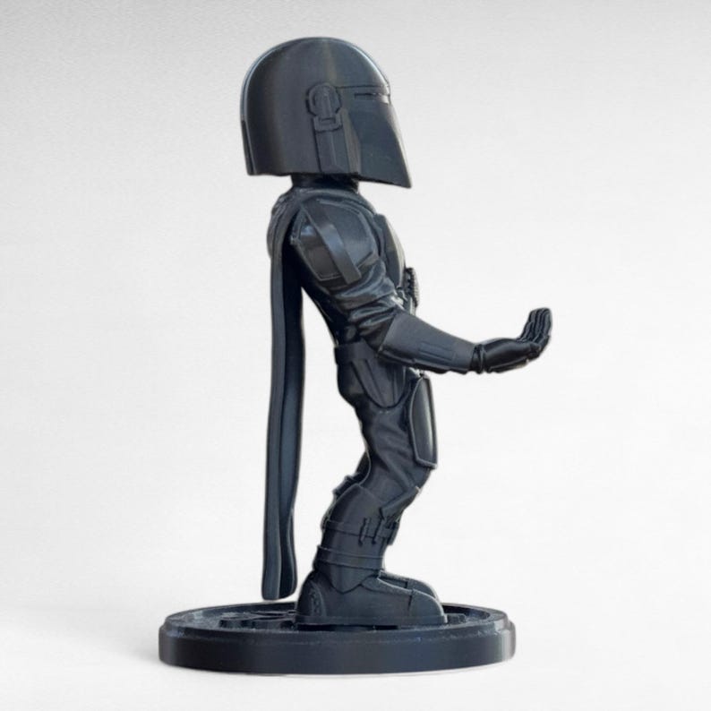 Pode incluir: Figura de a&ccedil;&atilde;o preta de um Mandaloriano, em p&eacute; sobre uma base redonda. A figura est&aacute; em uma pose din&acirc;mica, com um bra&ccedil;o estendido. O capacete e a armadura s&atilde;o detalhados, e uma capa pende nas costas. Feito de material escuro.