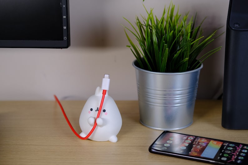 May include: A white ceramic cable holder shaped like a cartoon animal with a red cable coming out of its mouth. The cable holder is on a wooden surface next to a black smartphone and a potted plant.