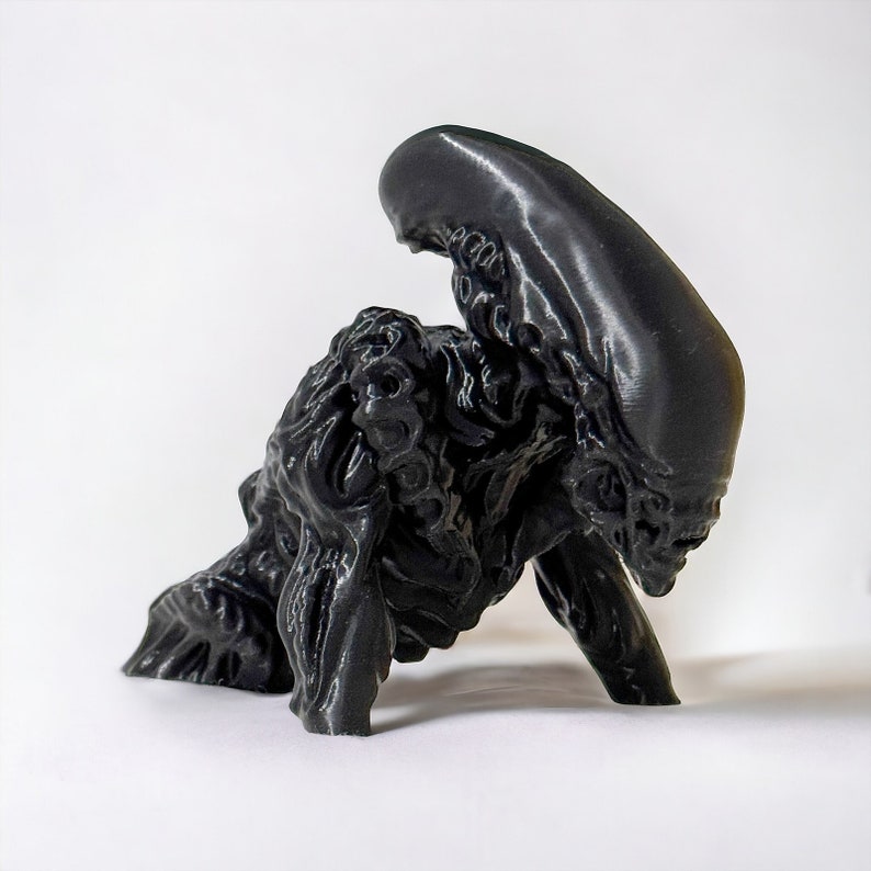 May include: A black 3D printed figurine of an alien creature with a large head and a long, thin body. The creature is in a crouched position, with its head tilted down.