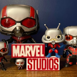 K&ouml;nnte beinhalten: Eine Sammlung von f&uuml;nf Ant-Man Funko Pop Figuren, darunter eine gro&szlig;e Ant-Man Figur, eine kleinere Ant-Man Figur und drei Ant-Man Figuren mit verschiedenen Helmen. Die Figuren stehen auf einer Holzoberfl&auml;che vor einem roten und wei&szlig;en Schild mit der Aufschrift "MARVEL STUDIOS".