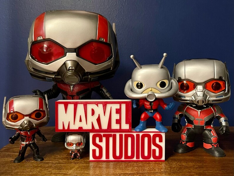 K&ouml;nnte beinhalten: Eine Sammlung von f&uuml;nf Ant-Man Funko Pop Figuren, darunter eine gro&szlig;e Ant-Man Figur, eine kleinere Ant-Man Figur und drei Ant-Man Figuren mit verschiedenen Helmen. Die Figuren stehen auf einer Holzoberfl&auml;che vor einem roten und wei&szlig;en Schild mit der Aufschrift "MARVEL STUDIOS".