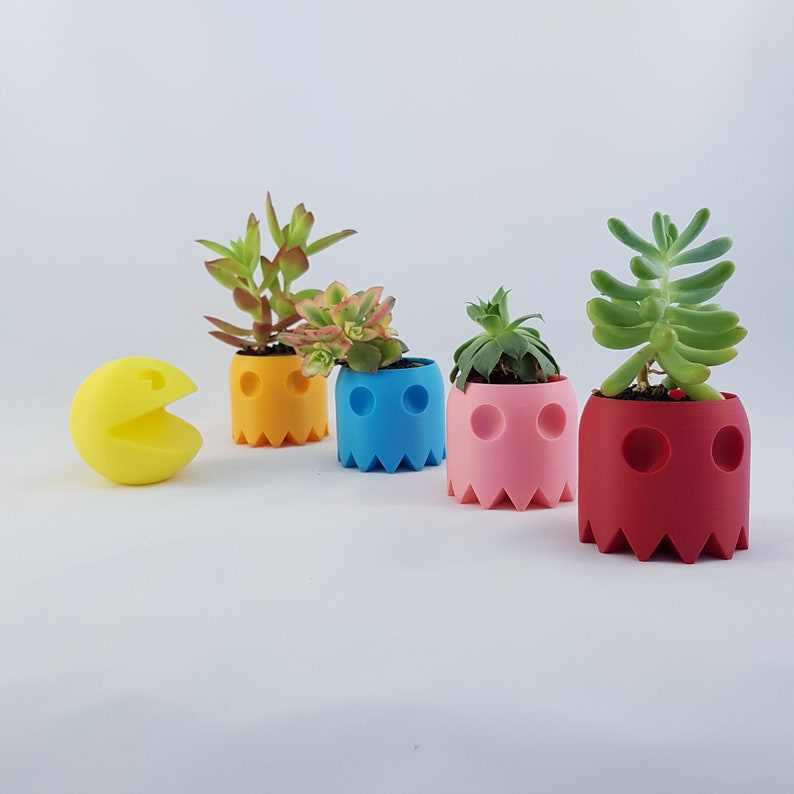 May include: Four colorful ghost-shaped planters with succulents. The planters are yellow, blue, pink, and red. A yellow Pac-Man figure is in the foreground.