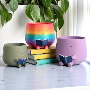 K&ouml;nnte beinhalten: Drei l&auml;chelnde Blument&ouml;pfe in Olivgr&uuml;n, Lavendel und Regenbogenfarben, die jeweils ein blaues Buch halten. Der Regenbogen-Topf steht auf einem Stapel B&uuml;cher. Diese skurrilen T&ouml;pfe sind ideal f&uuml;r kleine Pflanzen.