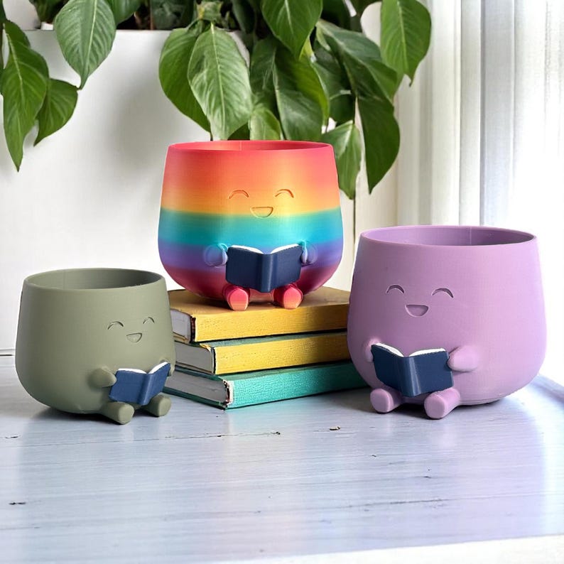 K&ouml;nnte beinhalten: Drei l&auml;chelnde Blument&ouml;pfe in Olivgr&uuml;n, Lavendel und Regenbogenfarben, die jeweils ein blaues Buch halten. Der Regenbogen-Topf steht auf einem Stapel B&uuml;cher. Diese skurrilen T&ouml;pfe sind ideal f&uuml;r kleine Pflanzen.
