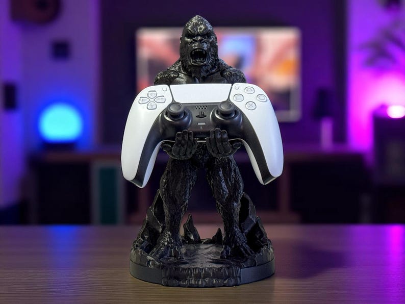 May include: A black gorilla statue holding a white and black video game controller. The statue has a detailed texture and is set on a round base. The controller has a sleek design with multiple buttons.