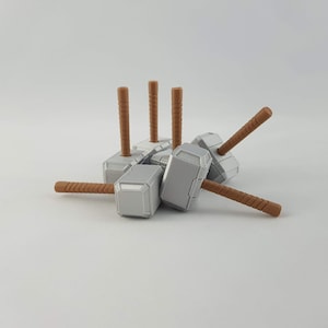 May include: Five miniature silver-colored Thor's hammers with brown wooden handles. The hammers are arranged in a pile with the handles pointing upwards.
