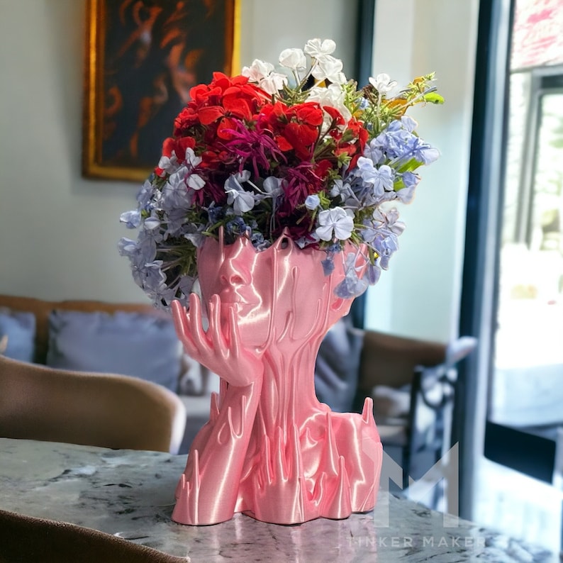 May include: A pink, abstract, 3D printed vase shaped like a human head with flowers in it. The vase is made of a smooth, glossy material and has a flowing, organic design. The flowers are a mix of red, white, and blue.