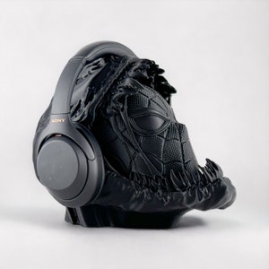 May include: Black headphones with a black and gray 3D printed stand shaped like a Venom character from Marvel Comics. The headphones have the word "SONY" printed on the side.