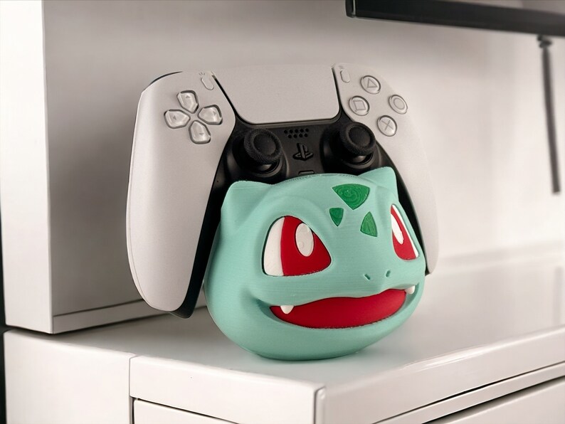 May include: A white and black PlayStation 5 controller is resting on a green and red Bulbasaur Pokemon character stand.