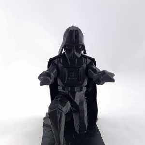 May include: A black 3D-printed figurine of Darth Vader, kneeling with his arms outstretched. The figure has a detailed helmet, armor, and cape. The figurine is on a black base, set against a white background.