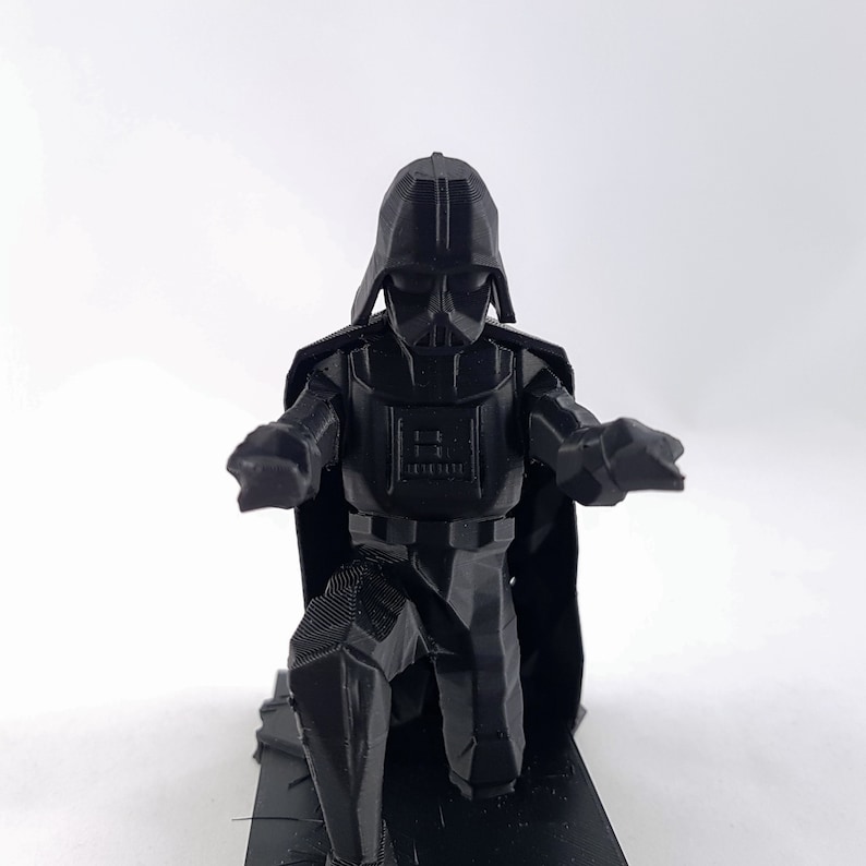 May include: A black 3D-printed figurine of Darth Vader, kneeling with his arms outstretched. The figure has a detailed helmet, armor, and cape. The figurine is on a black base, set against a white background.