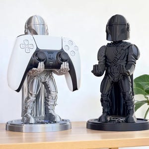 Pode incluir: Dois suportes de comando com tema Mandalorian. Um suporte prateado segura um comando PlayStation 5 branco e preto. O outro &eacute; uma figura preta de Mandalorian. Ambos os suportes est&atilde;o em bases redondas e s&atilde;o exibidos em uma superf&iacute;cie de madeira.