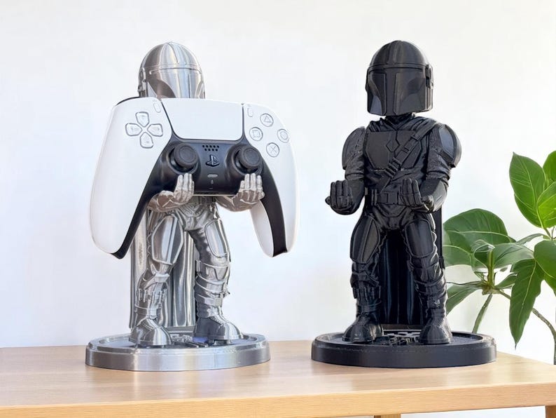 Pode incluir: Dois suportes de comando com tema Mandalorian. Um suporte prateado segura um comando PlayStation 5 branco e preto. O outro &eacute; uma figura preta de Mandalorian. Ambos os suportes est&atilde;o em bases redondas e s&atilde;o exibidos em uma superf&iacute;cie de madeira.
