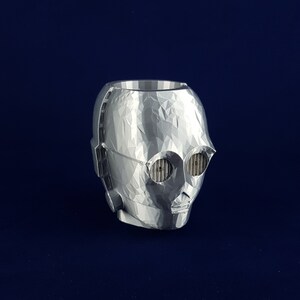 May include: A silver 3D printed planter shaped like the head of C-3PO from Star Wars. The planter has a textured surface and features the character's signature eyes and mouth.