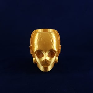 May include: A gold-coloured 3D printed planter shaped like a robot head. The planter has a geometric design and features two small, rectangular holes for eyes.