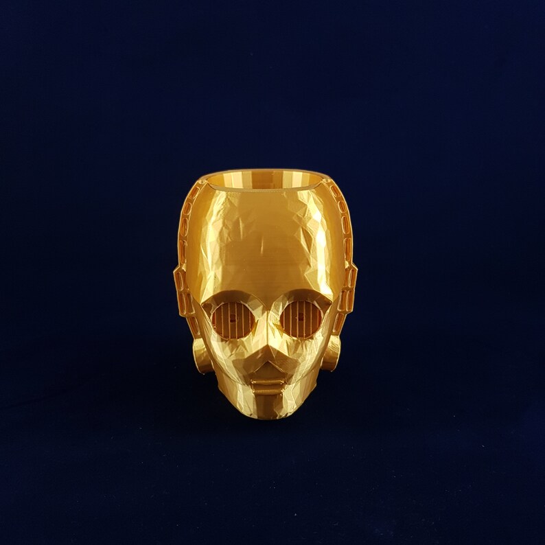 May include: A gold-coloured 3D printed planter shaped like a robot head. The planter has a geometric design and features two small, rectangular holes for eyes.