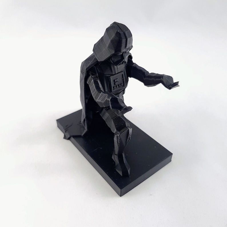 May include: A black 3D-printed Darth Vader figurine in a kneeling pose. The figurine is on a rectangular base and features detailed geometric facets. The figure is a representation of the Star Wars character.