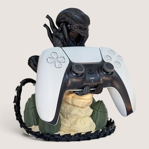 Peut inclure: Une figurine noire de Xénomorphe servant de support de manette, tenant une manette de jeu PlayStation 5 blanche et noire. La base présente une surface texturée beige rappelant la roche, entourée d'œufs verts et d'une longue queue noire.