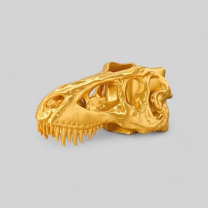 Op de afbeelding: Een gouden dinosaurus schedel sculptuur. De schedel is gedetailleerd met tanden en oogkassen.
