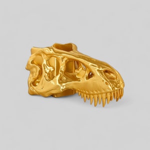 Op de afbeelding: Een gouden dinosaurus schedel sculptuur. De schedel is gedetailleerd met tanden en oogkassen.