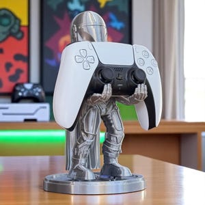 Pode incluir: Um suporte para comando PlayStation 5 com tema Mandalorian, na cor prateada. O suporte tem a forma da personagem Mandalorian, segurando um comando PlayStation 5 branco e preto. A base &eacute; circular e prateada.