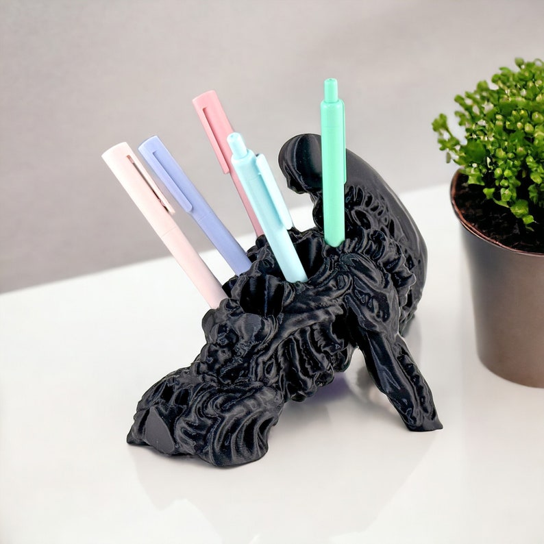 May include: A black 3D printed pen holder shaped like a creature with a textured surface. The holder has four slots for pens and is holding four pens: one pink, one light blue, one blue, and one green.