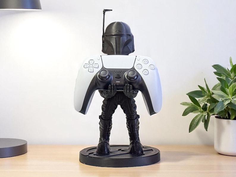 May include: A black and white Mandalorian-themed controller stand. The stand is shaped like the Mandalorian, holding a white and black PlayStation 5 controller. The stand is on a black circular base, perfect for gamers.