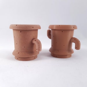 May include: Two terracotta-colored ceramic planters shaped like cacti with small holes throughout. The planter on the left has a single arm, while the planter on the right has two arms.