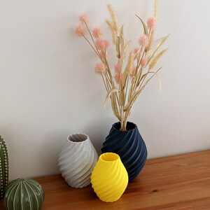 May include: Three vases of different colors and designs. A gray vase, a yellow vase, and a black vase. The vases are arranged on a wooden surface. Dried flowers are in the black vase.