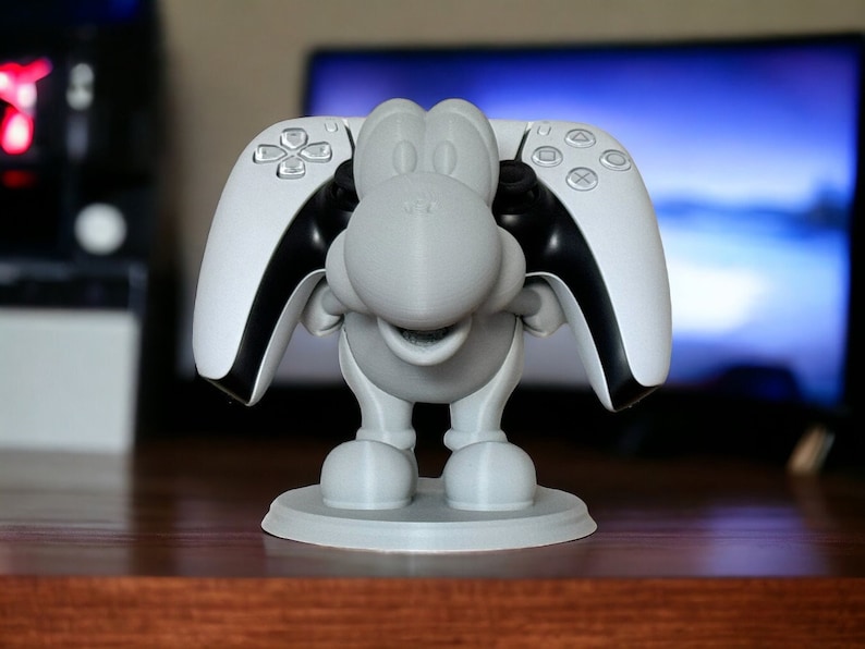 May include: A 3D printed gray figurine of a cartoon character holding two white and black video game controllers. The character has a wide smile and is standing on a round base.