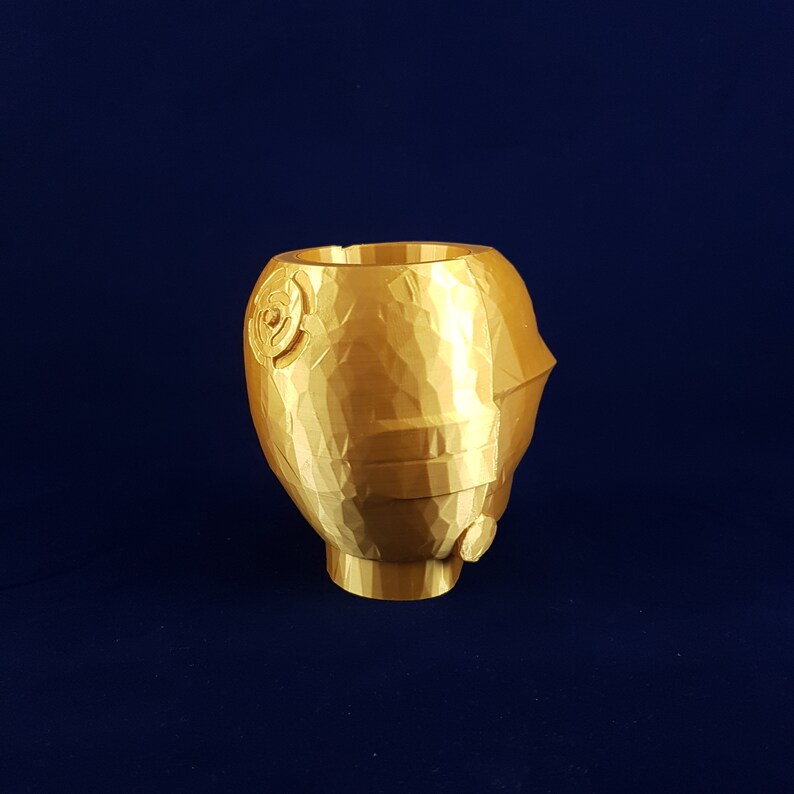 May include: A gold-coloured, 3D printed planter shaped like a stylised head. The planter has a textured surface and a small opening at the top for planting.