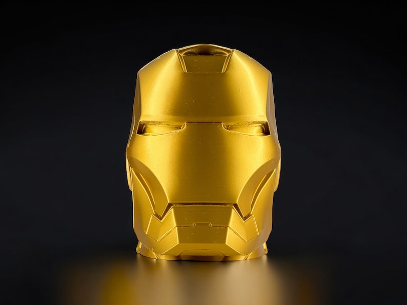 K&ouml;nnte beinhalten: Eine goldfarbene Metallskulptur eines Superheldenkopfes. Die Skulptur ist detailliert mit einer Maske und Gesichtsz&uuml;gen.