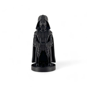 May include: A black plastic figurine of Darth Vader from Star Wars. The figurine is standing with its arms crossed and is wearing a black cape. The figurine is on a black base.